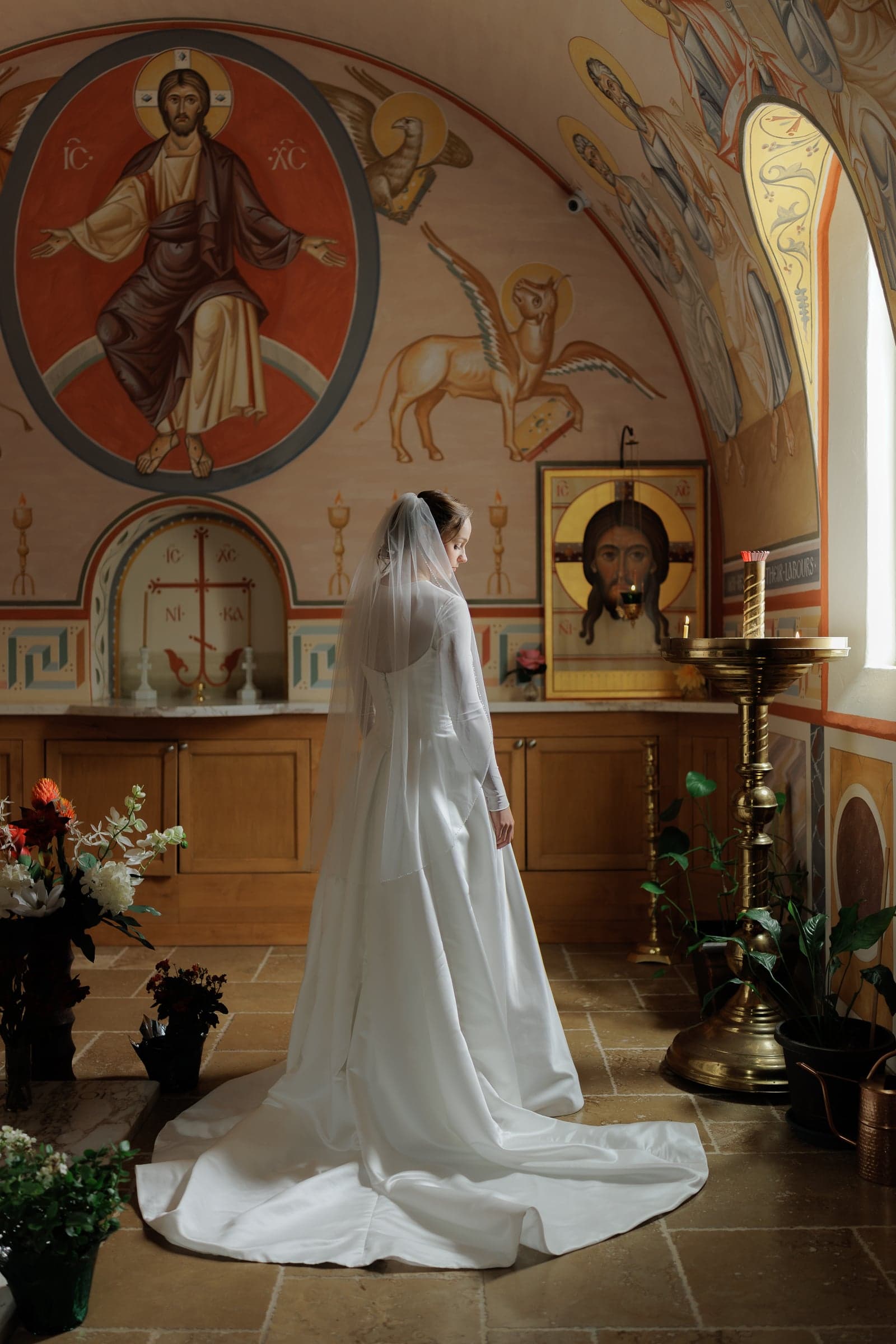 A veiled bride stands beside a candle stand before an icon of Christ in an Orthodox church