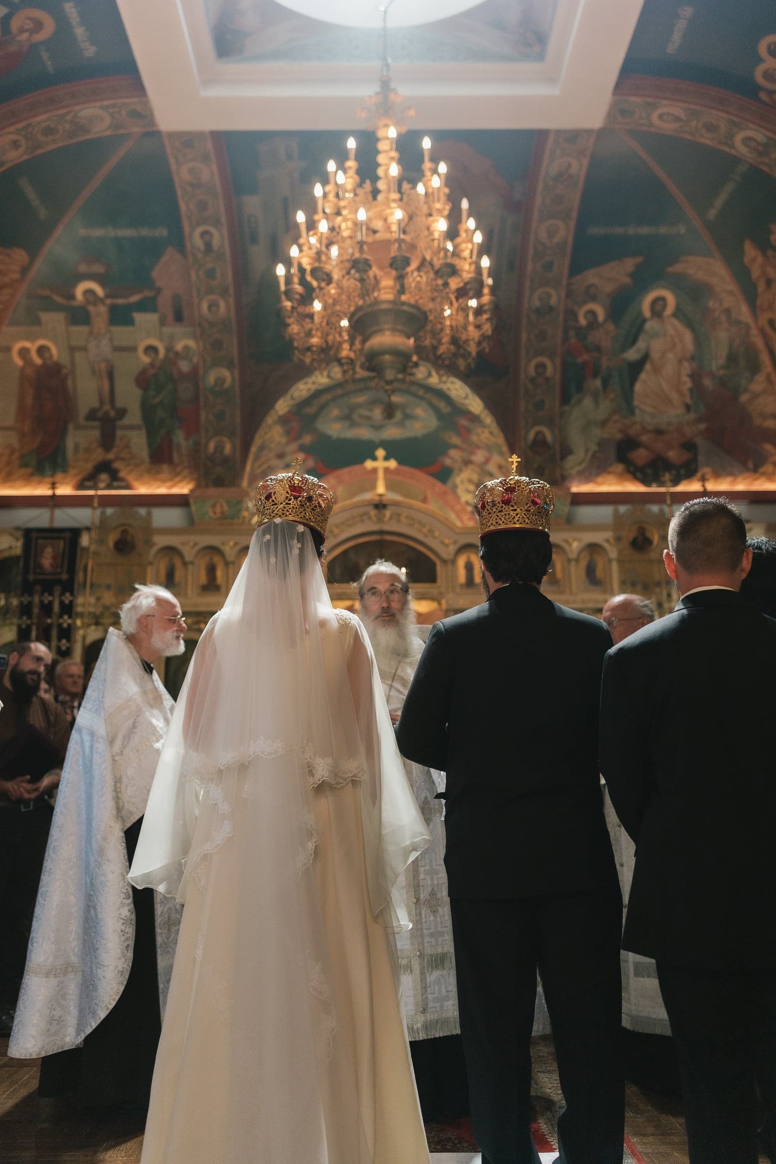 A crowned Orthodox bride and groom stand beneath a church chandelier during the wedding service