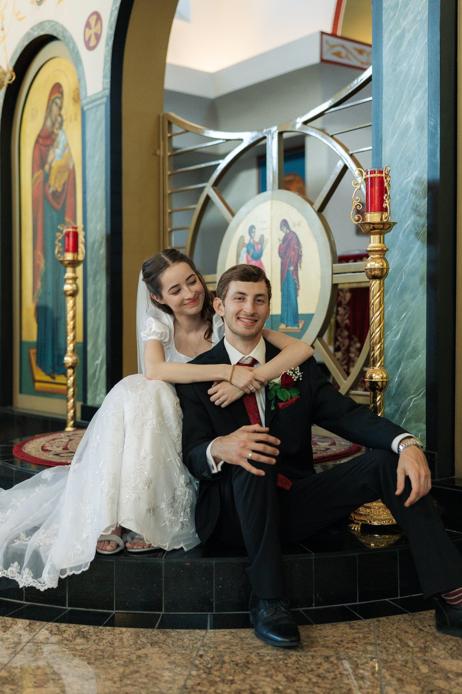 A bride sits behind the groom with her arms around his shoulders inside an Orthodox church